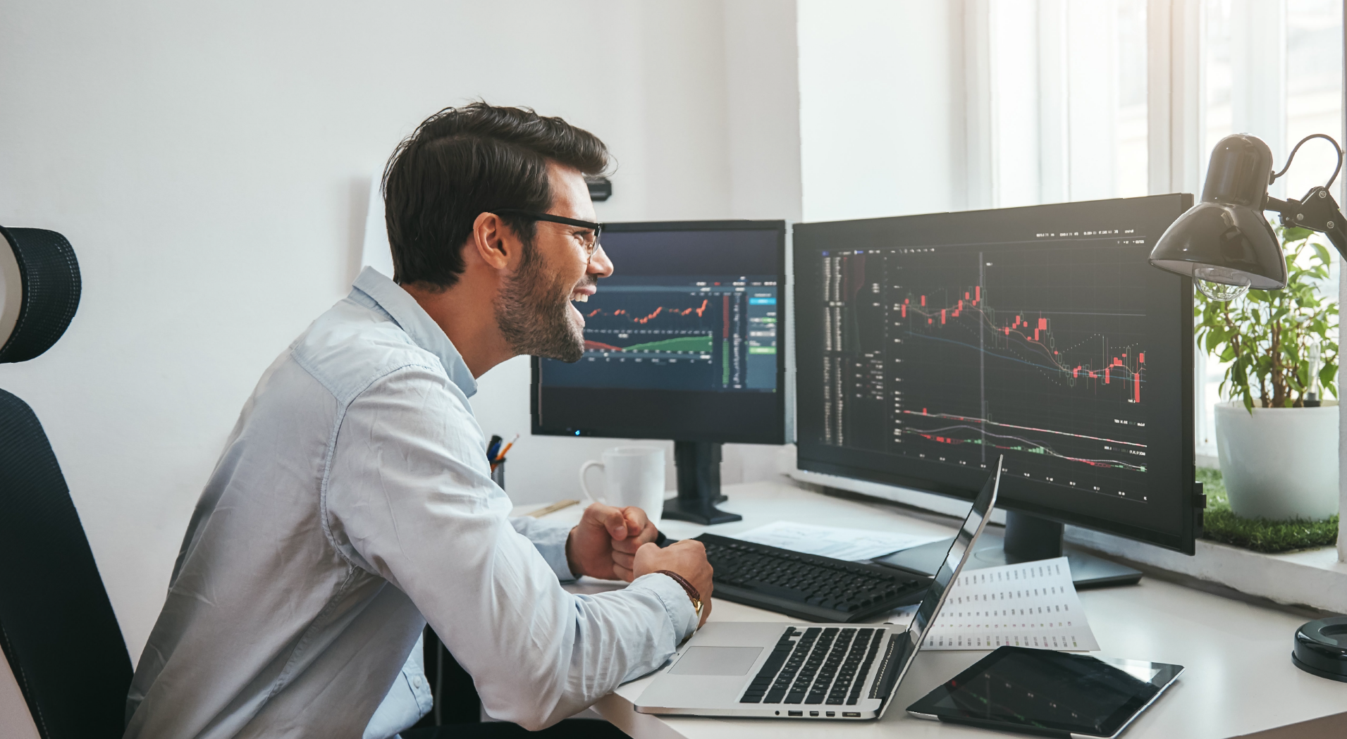 Man in glasses excitedly looking at financial charts on dual monitors at a desk with laptop and tablet.
