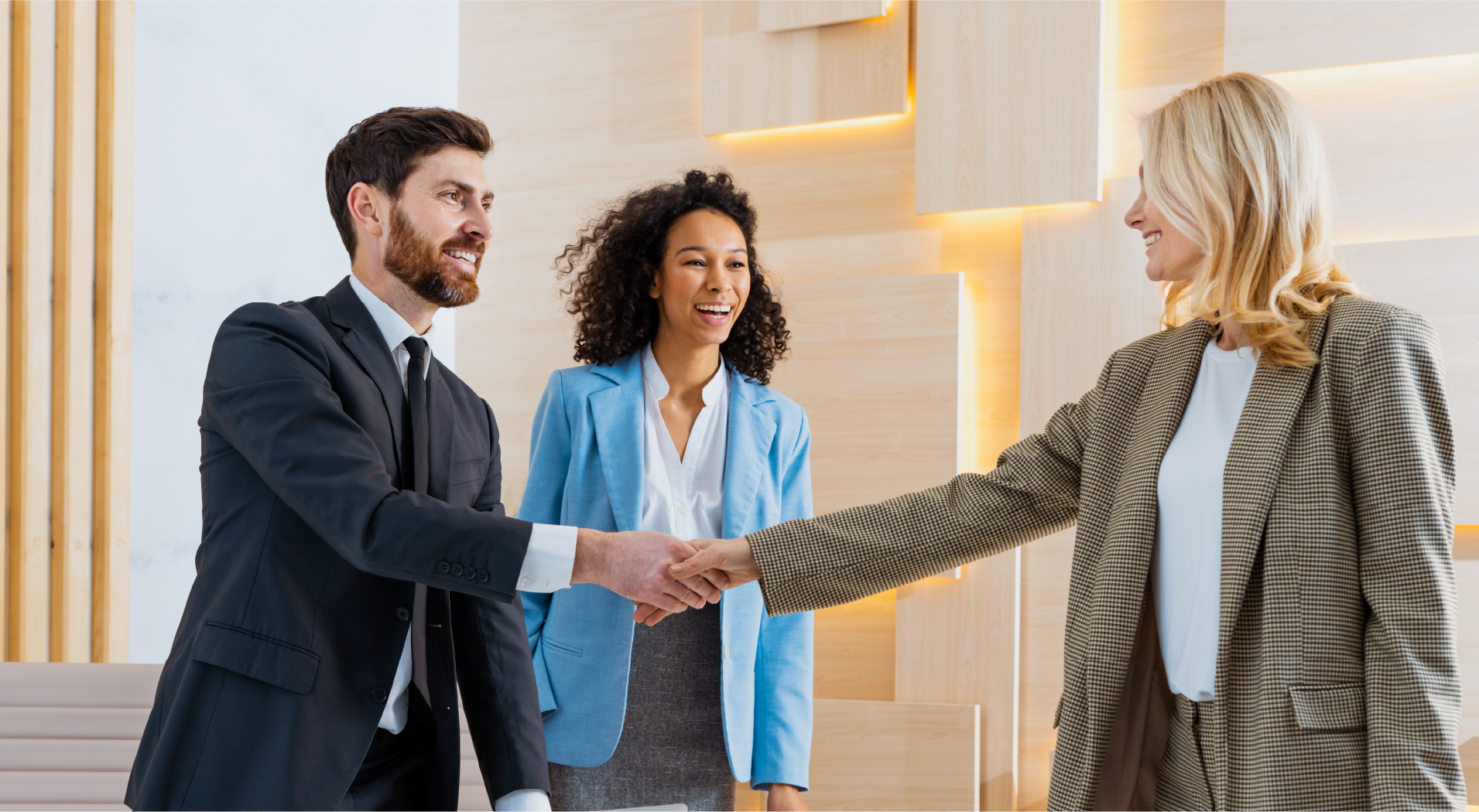 Two businesspeople shaking hands while a colleague in a blue blazer watches and smiles in a modern office.