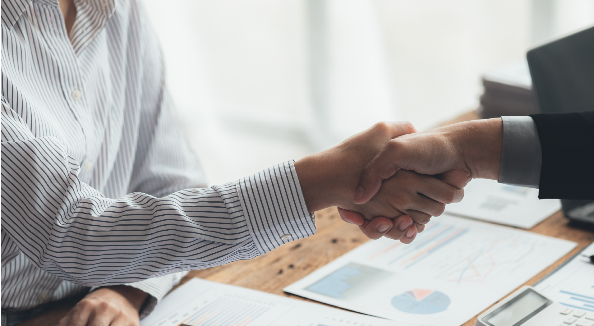 Two people shaking hands over a desk with business charts and documents.