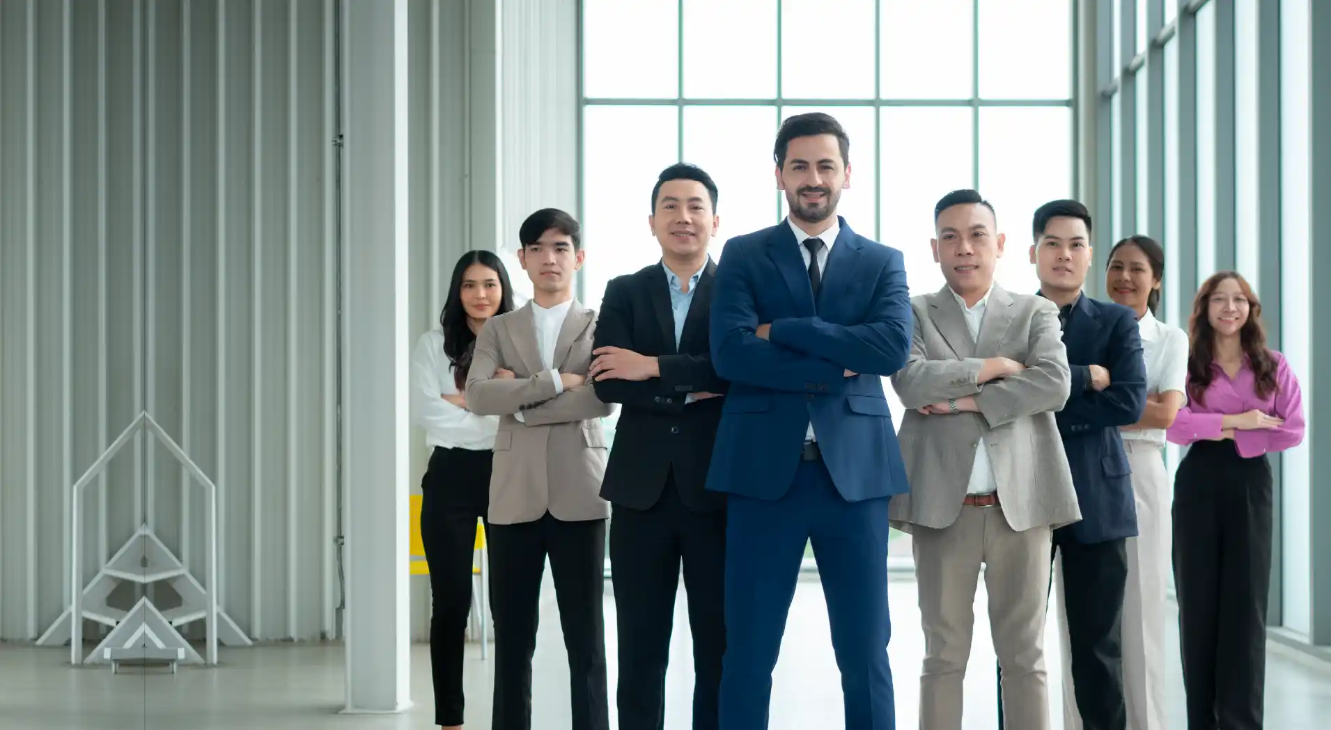 Confident business team of eight diverse professionals standing in a bright modern office with arms crossed.