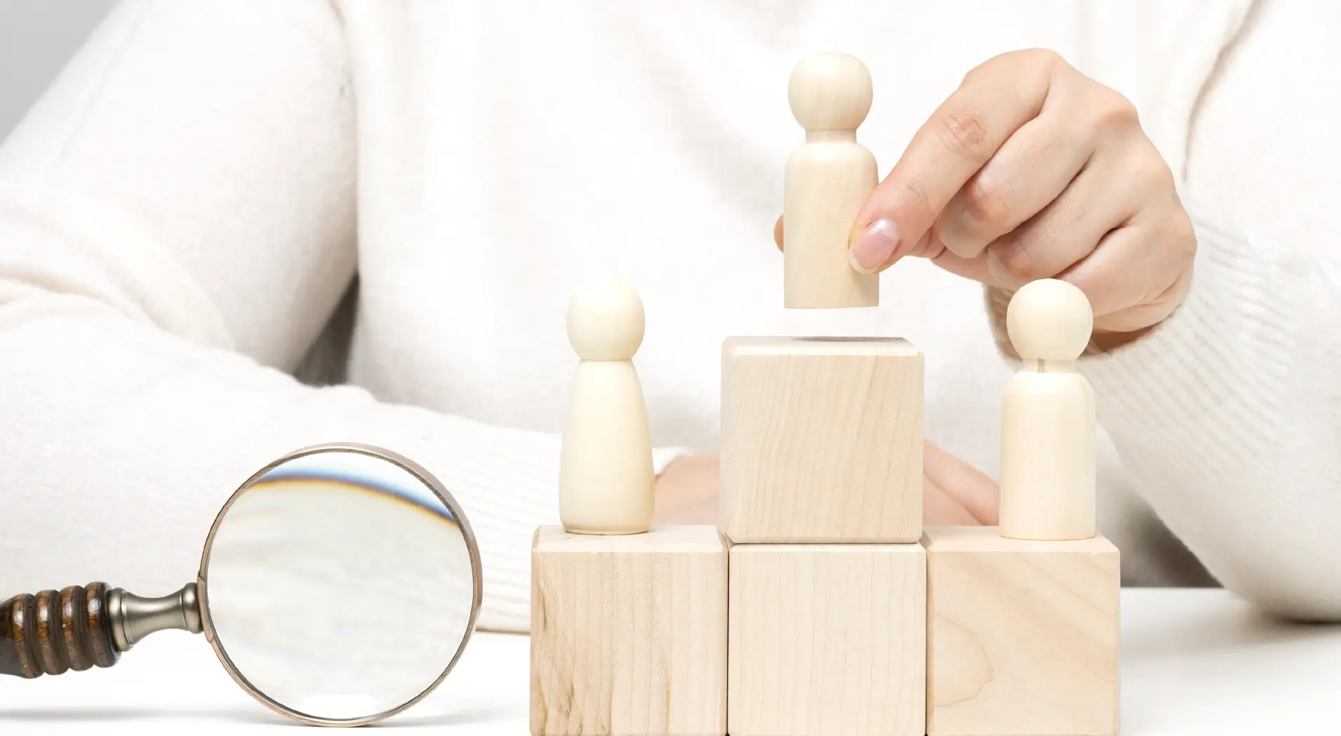 Hand placing a wooden figure on top of stacked wooden blocks with two other figures on lower blocks and a magnifying glass nearby.