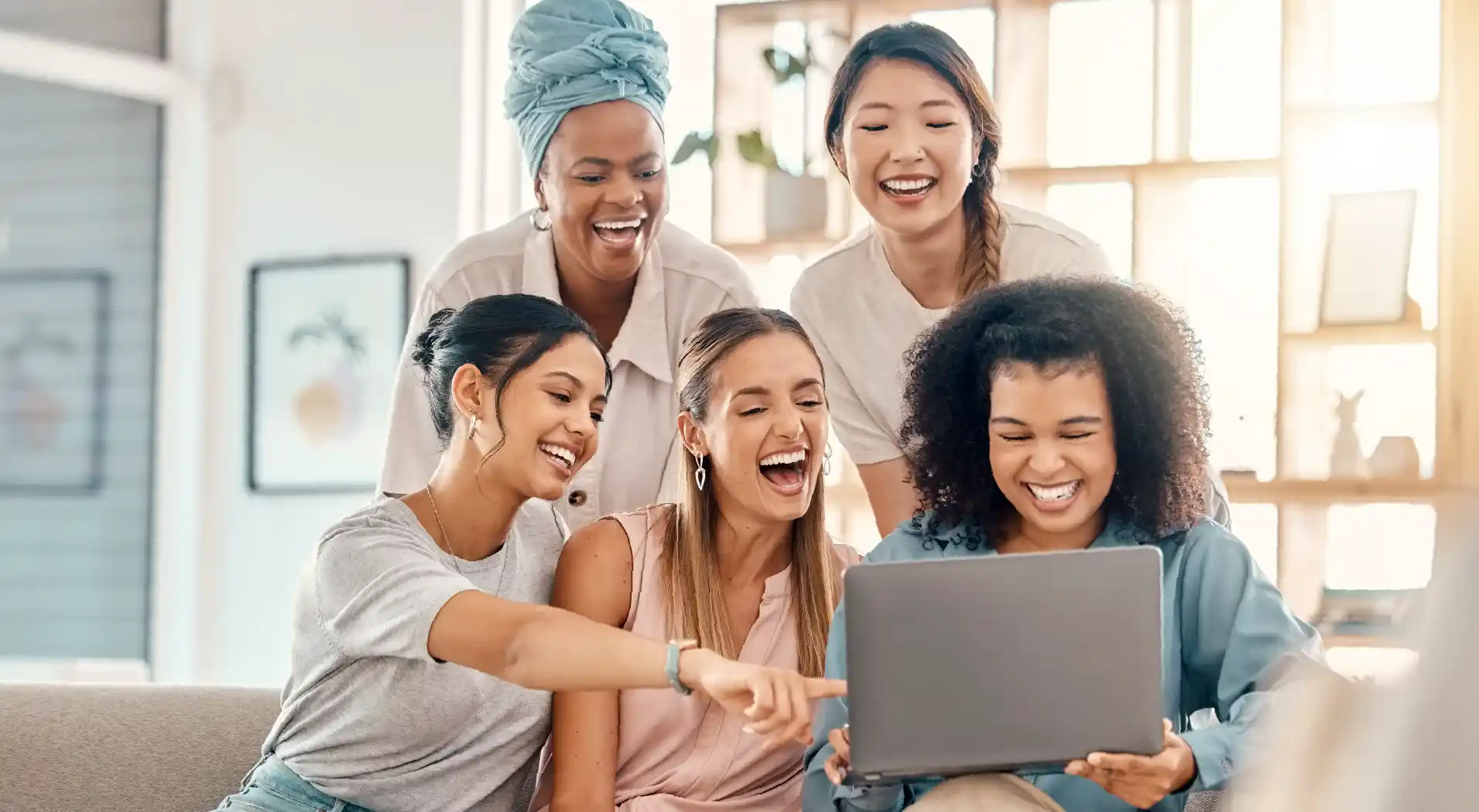 Group of five diverse young women smiling and laughing while looking at a laptop together in a bright room.