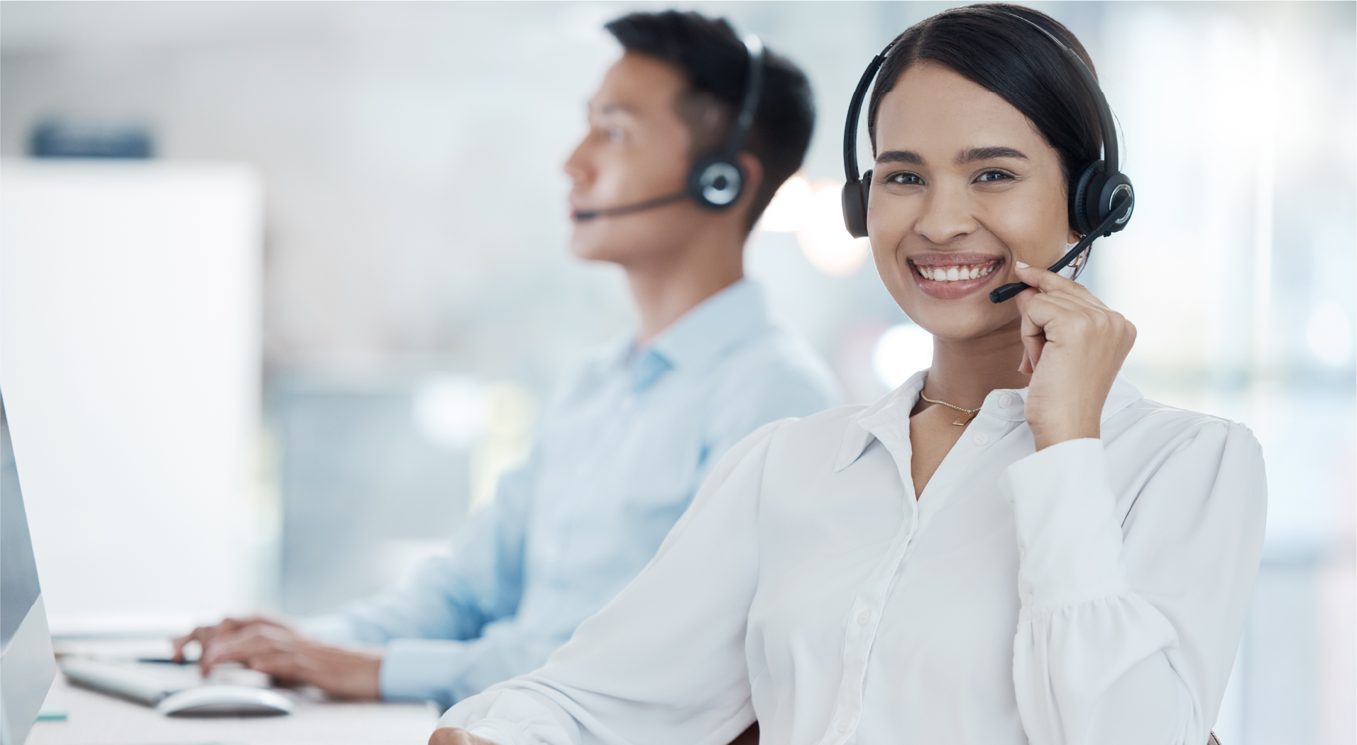 Smiling woman wearing a headset at a customer service desk with a man working in the background.