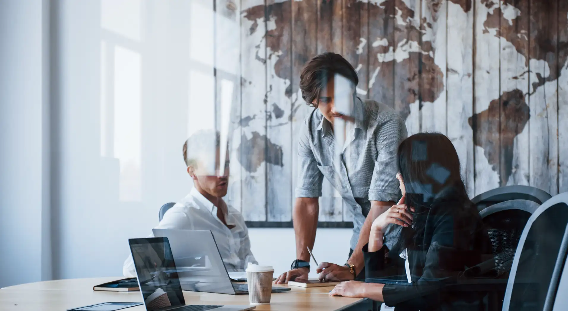 Three colleagues having a discussion around a table with laptops and a coffee cup, in a room with a world map on the wall.