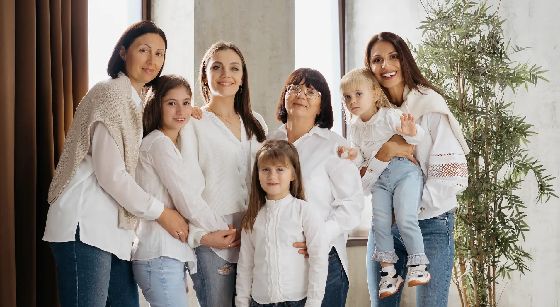 Seven women and girls of different ages standing together indoors wearing white tops and jeans, smiling at the camera.