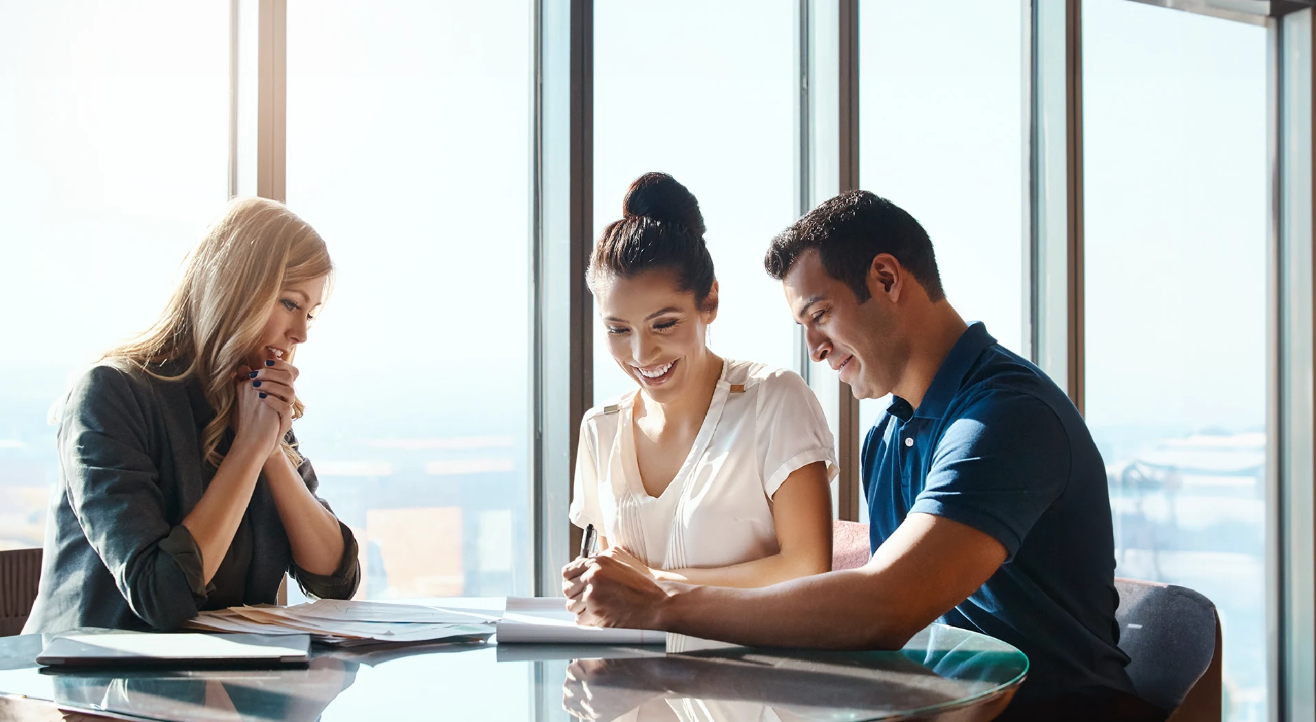 Two women and a man sitting at a glass table in a bright office, reviewing documents together with the man writing on paper.