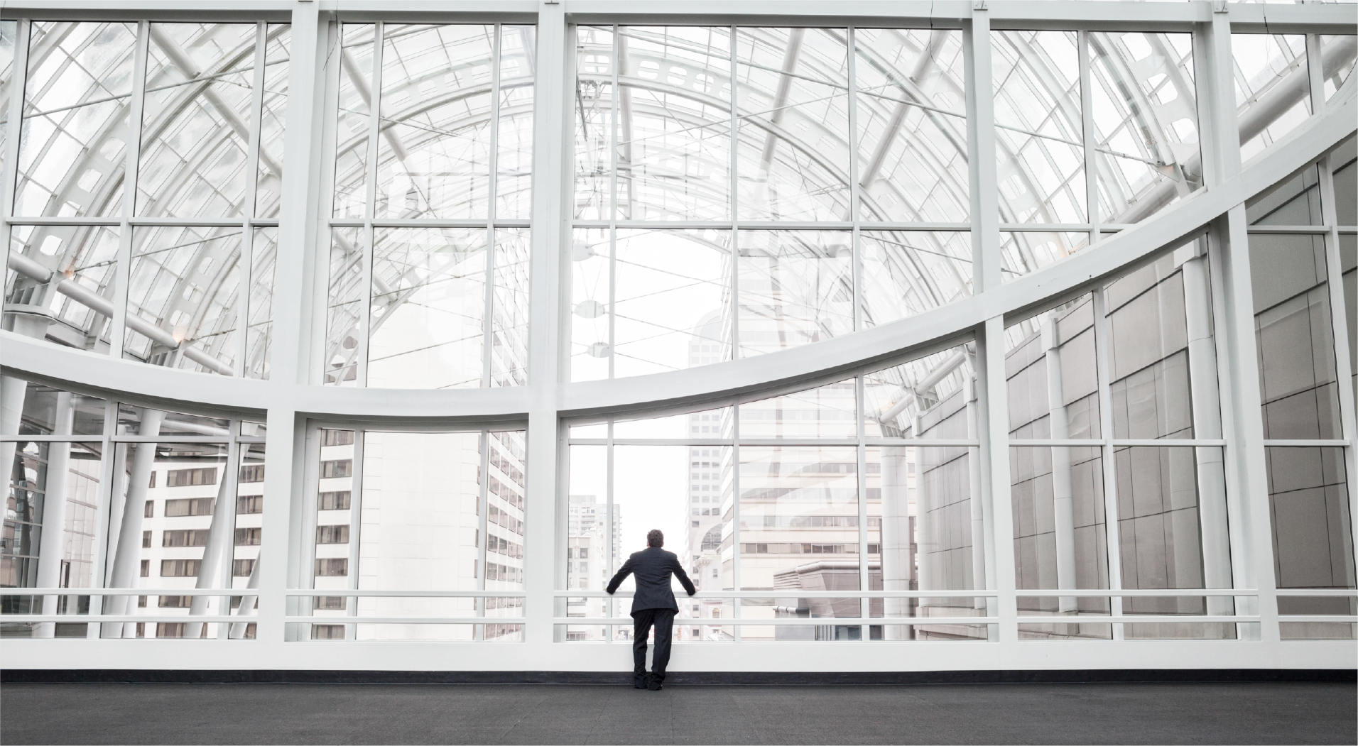 Man in business suit standing alone by large modern glass windows overlooking city buildings.
