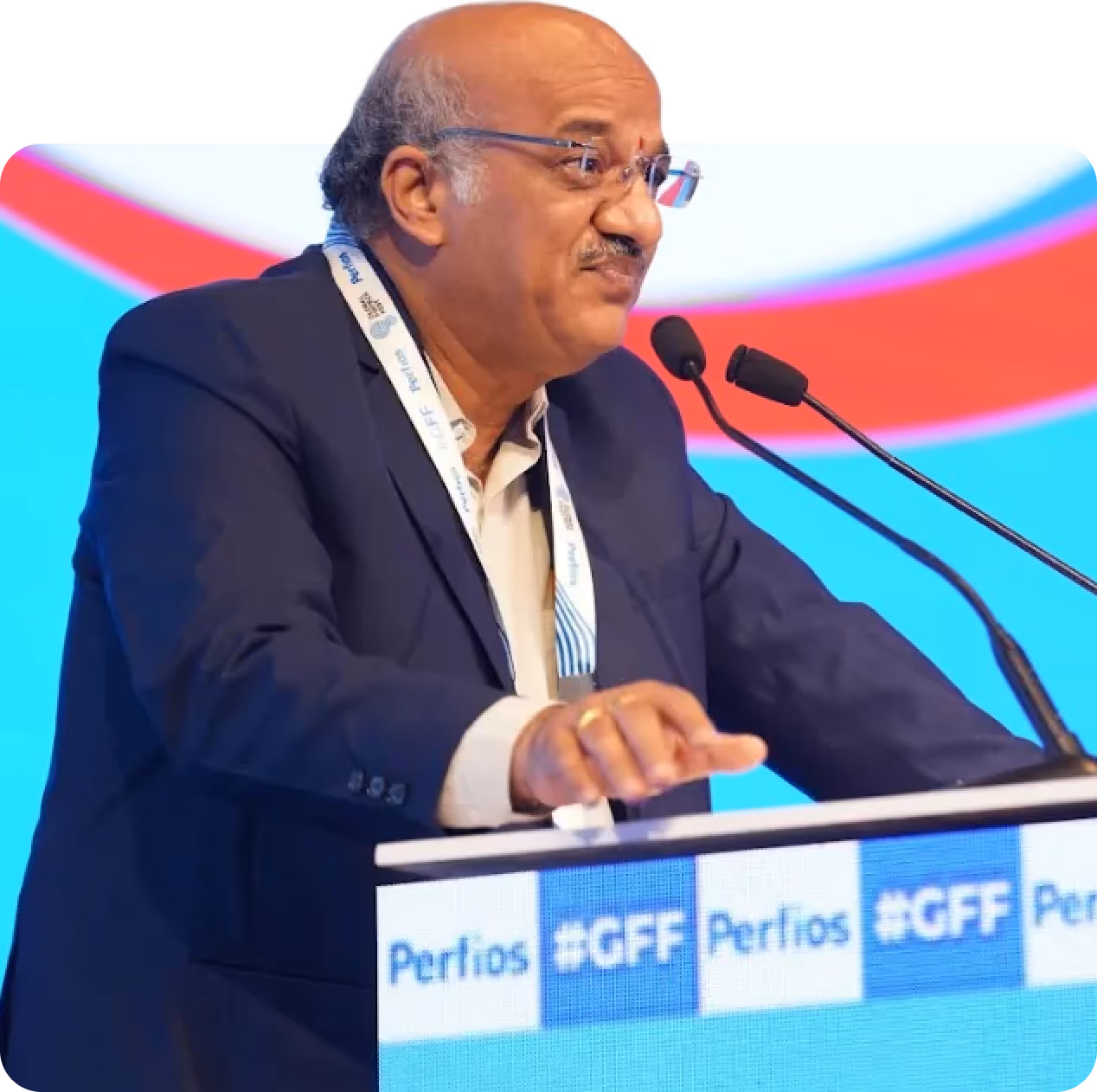 Man wearing glasses and a suit speaking at a podium with microphones and a blue and red background.