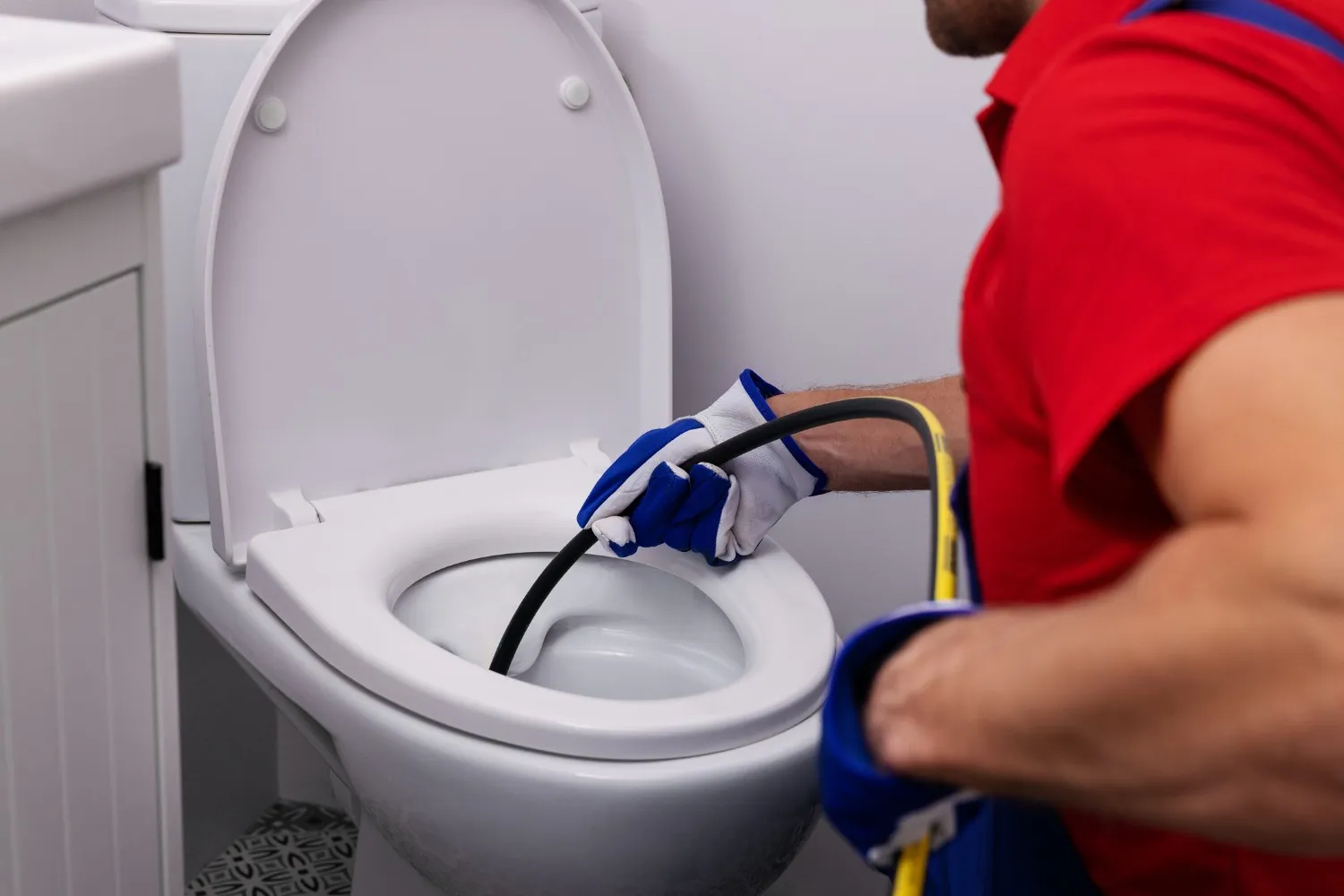 A plumber in a red shirt and blue gloves is using a hydro-jetting tool to unclog a toilet.