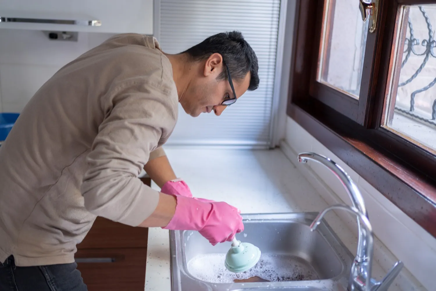 A male plumber in pink gloves and a beige hoodie is using a plunger to unclog a stainless steel kitchen sink.