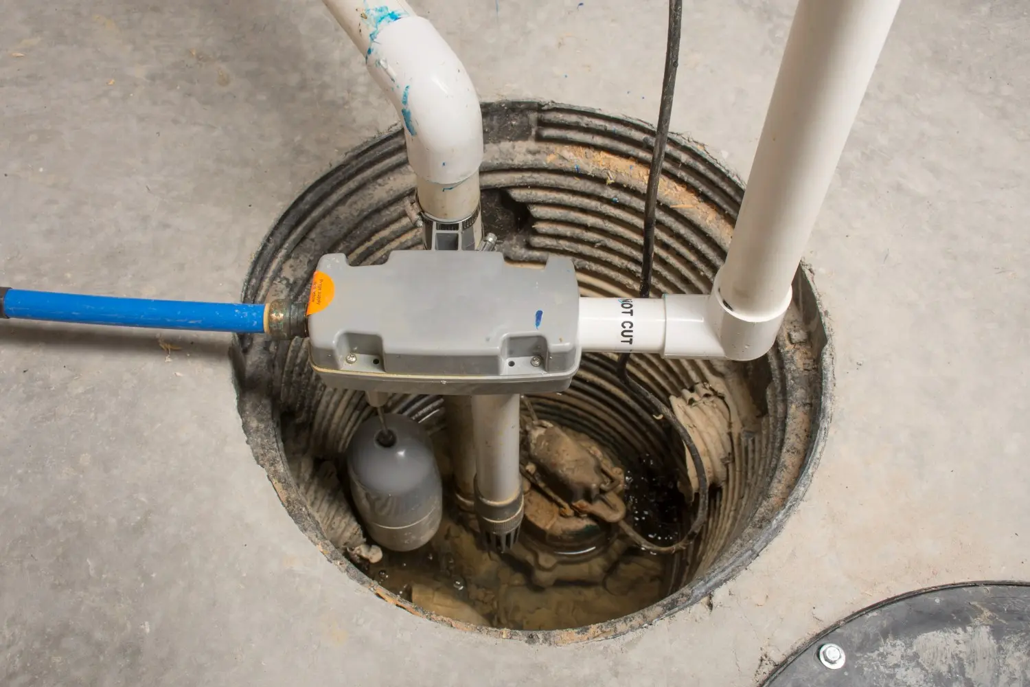 A top-down view of a sump pump installed in a basement pit, with various white and blue pipes leading to a gray control box and a floating gray sensor.