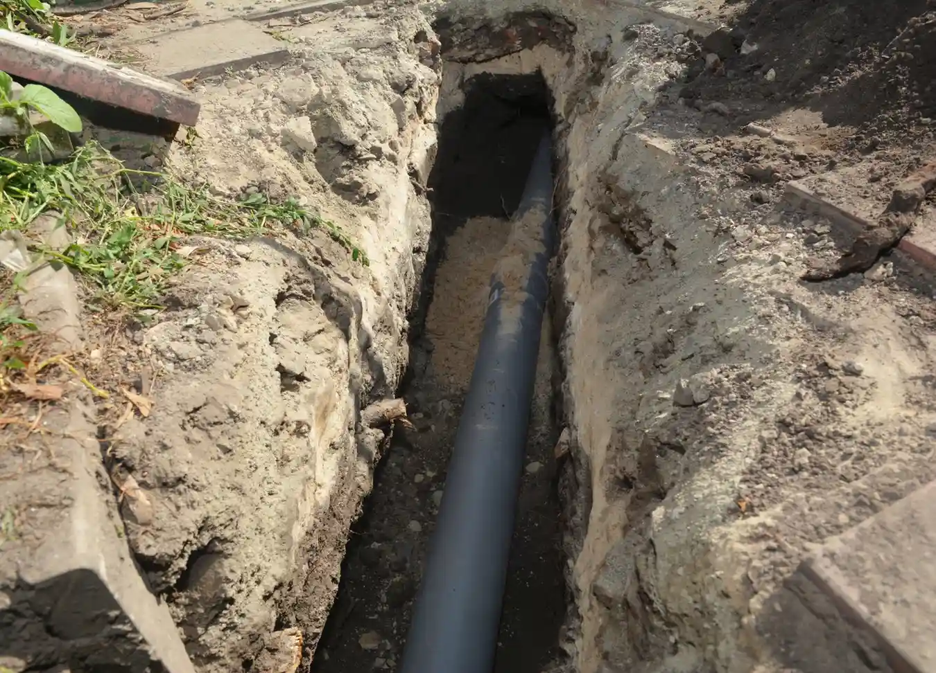 A large gray sewer pipe is shown in a narrow, deep trench dug in the ground with concrete borders.