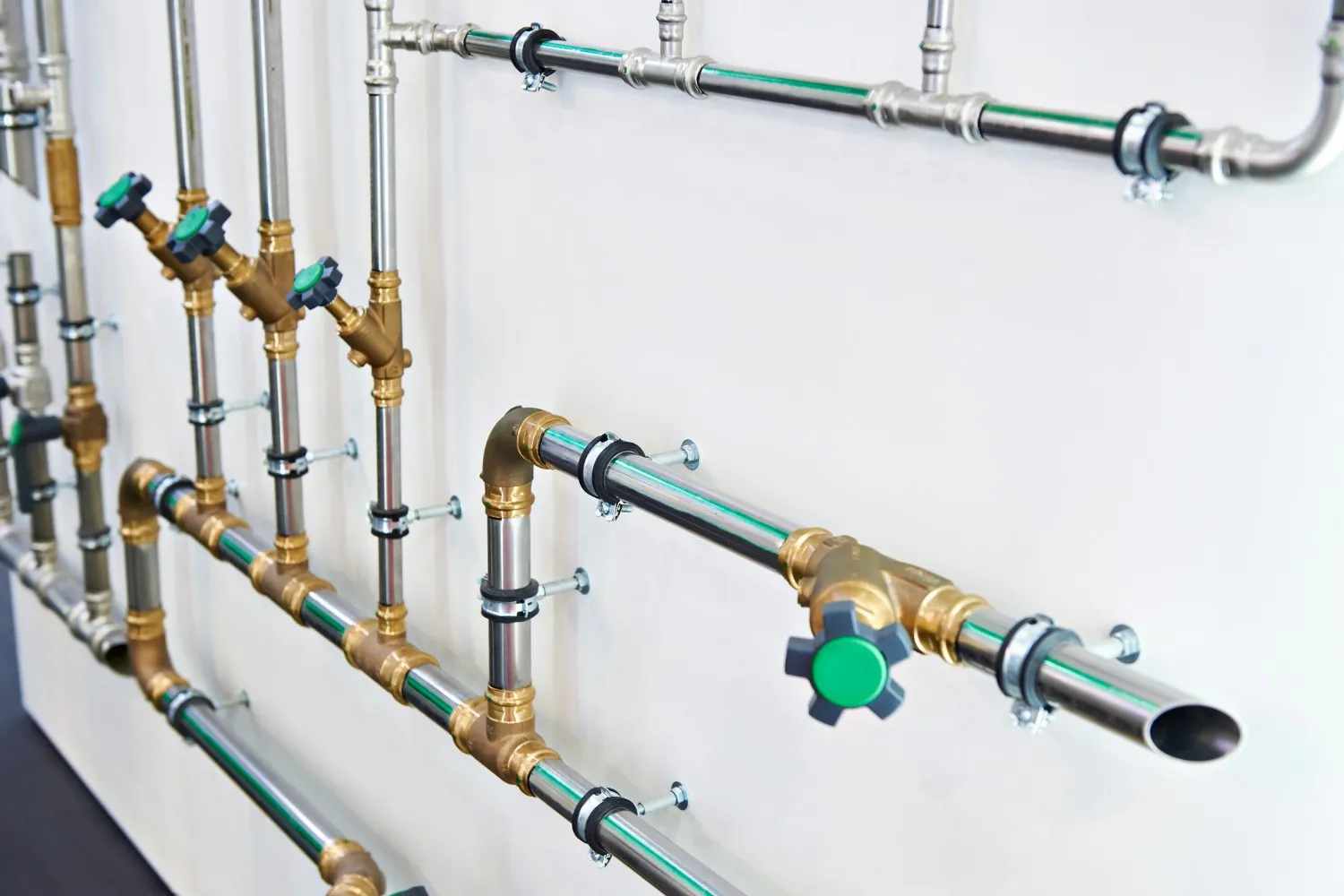 A clean and complex network of metal pipes, valves, and fittings is mounted on a white wall.