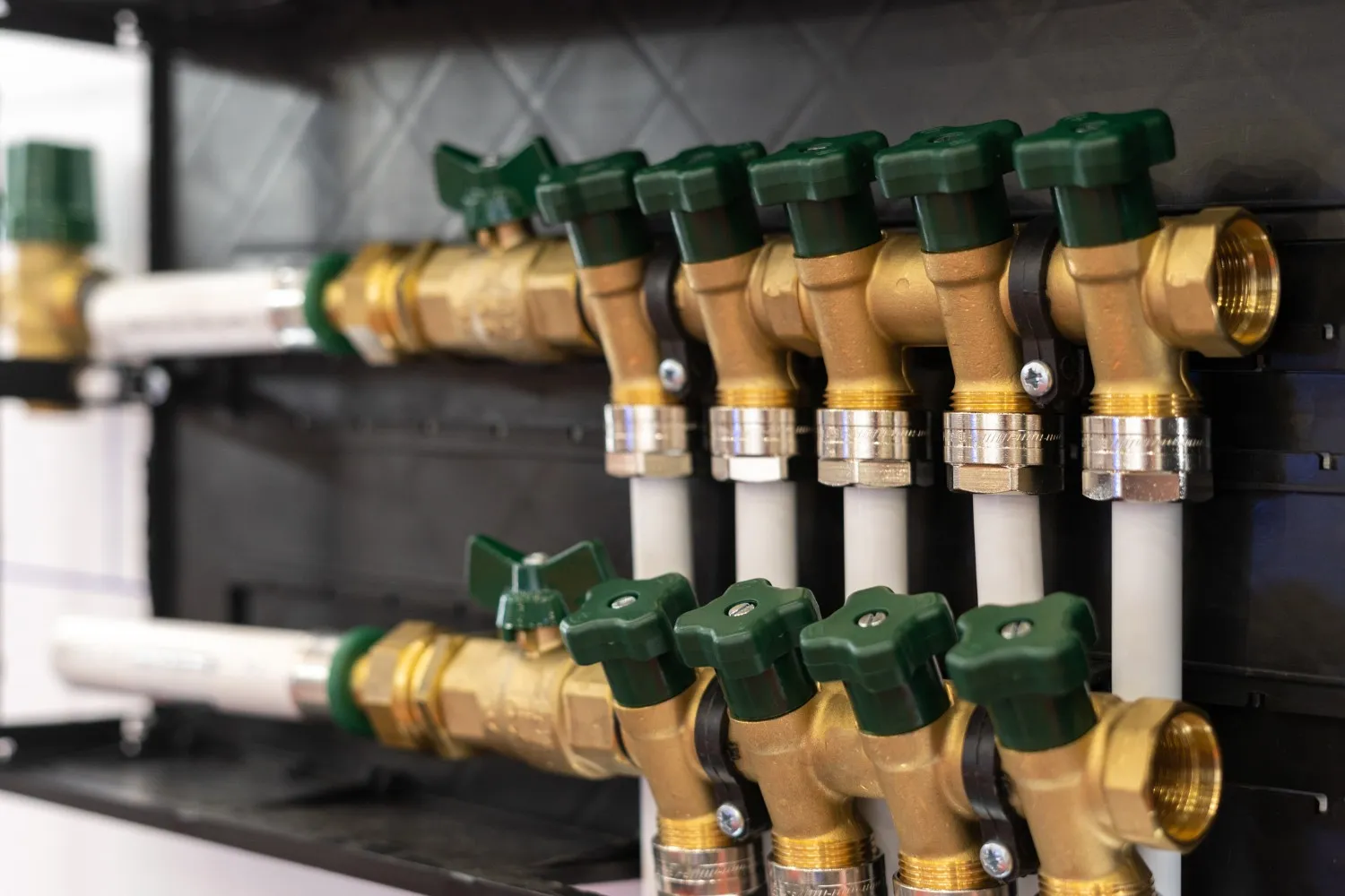 A close-up of a plumbing manifold shows a series of brass valves with green handles connected to white pipes.