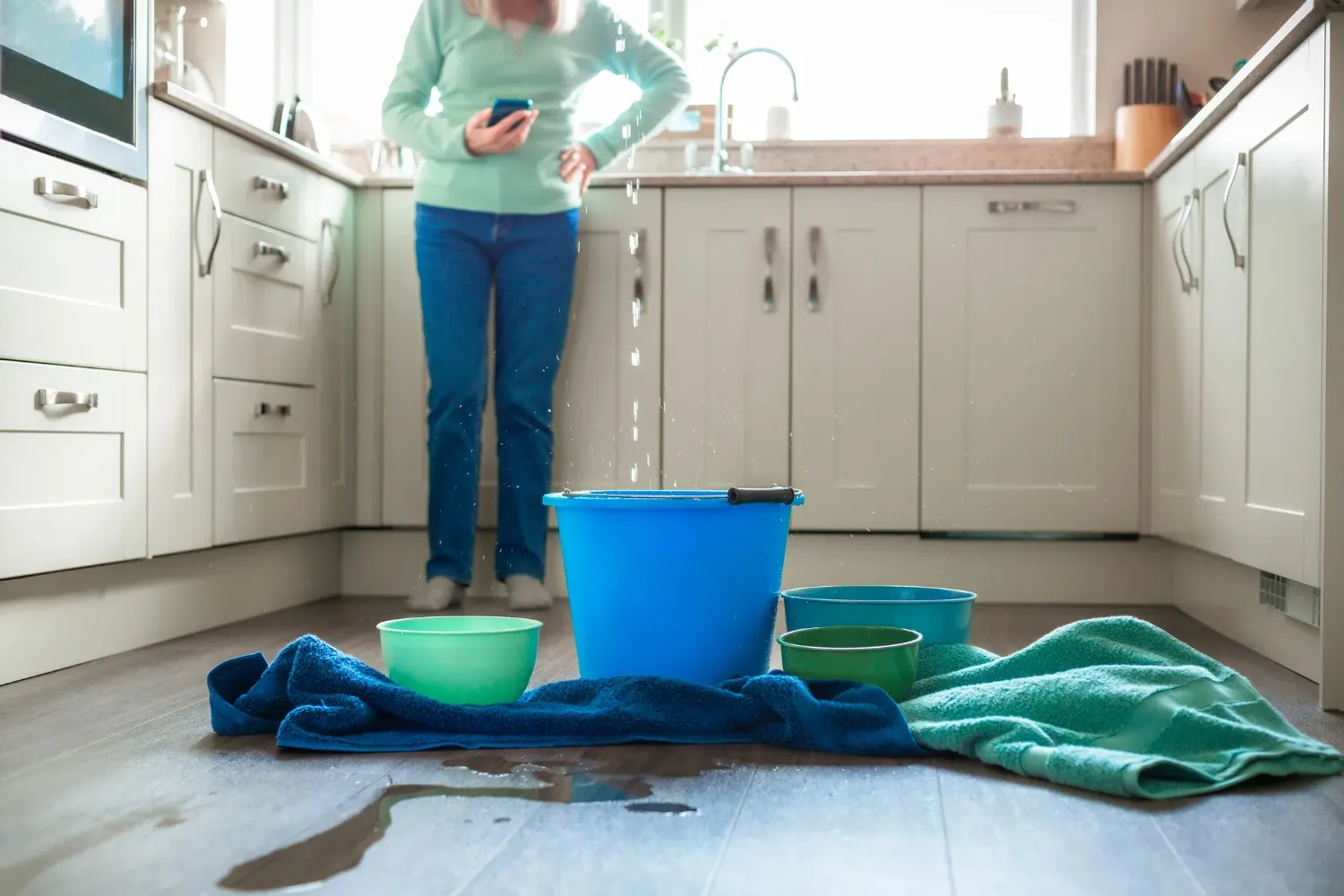 A worried woman is standing in her kitchen while a large leak is dripping into a blue bucket and several bowls on the floor.