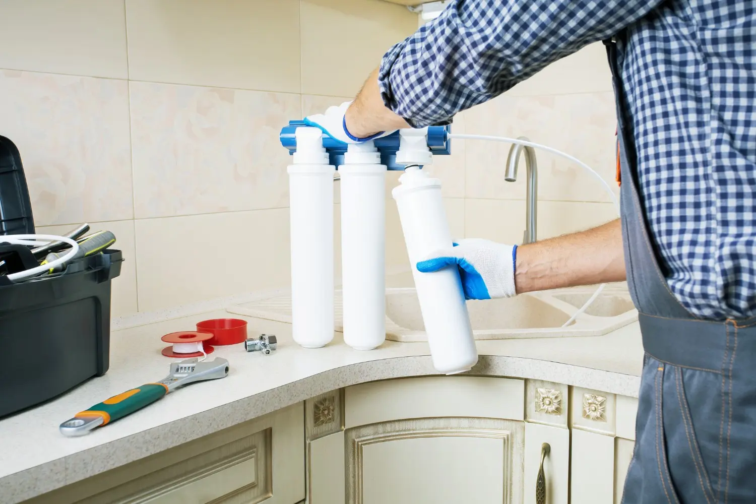 A plumber is wearing gloves and a checkered shirt while installing three white filter cartridges into a water filtration system under a kitchen sink.