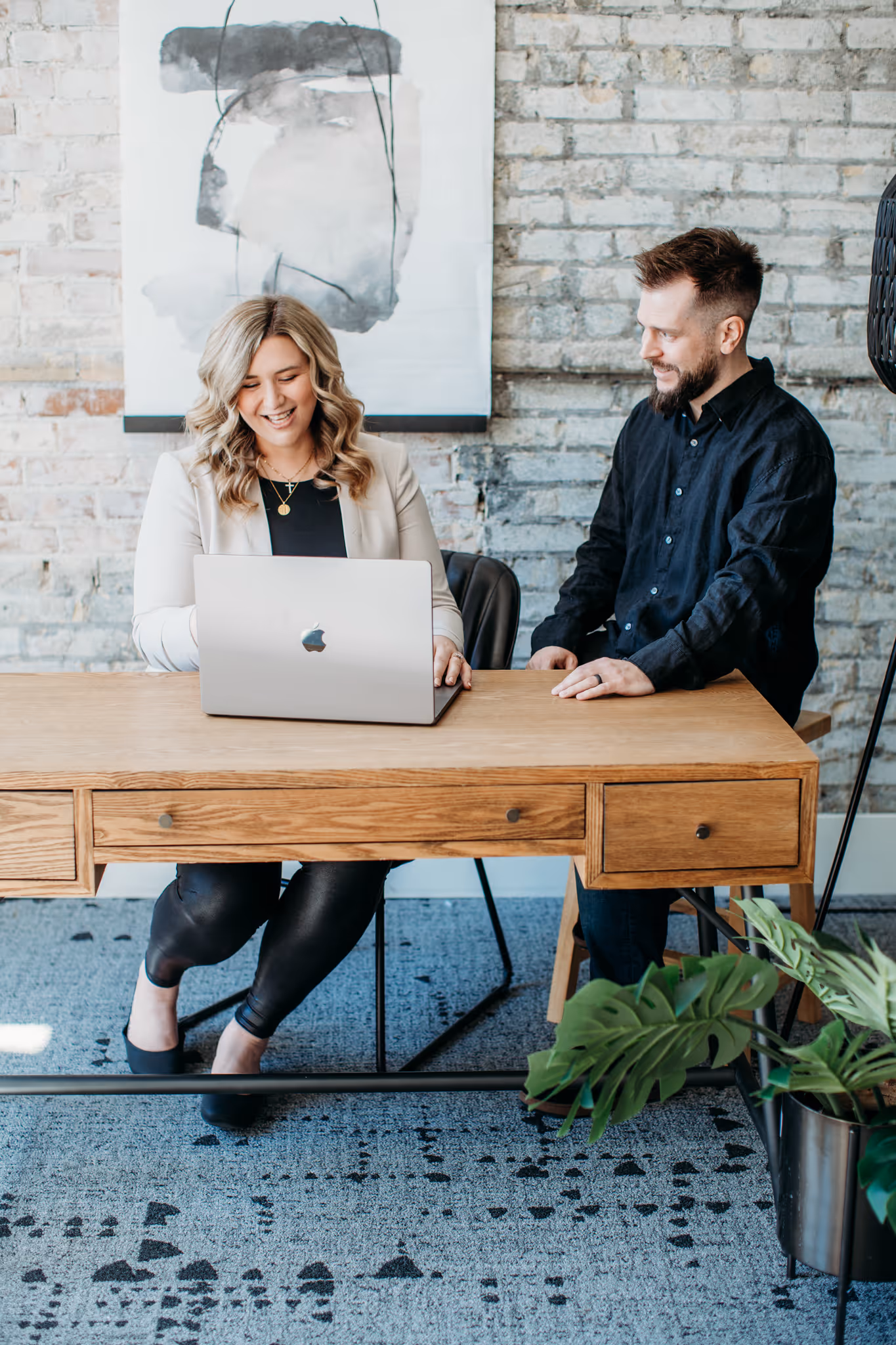 Smiling woman and man seated at a wooden desk with a laptop in a modern office with brick walls and abstract art.