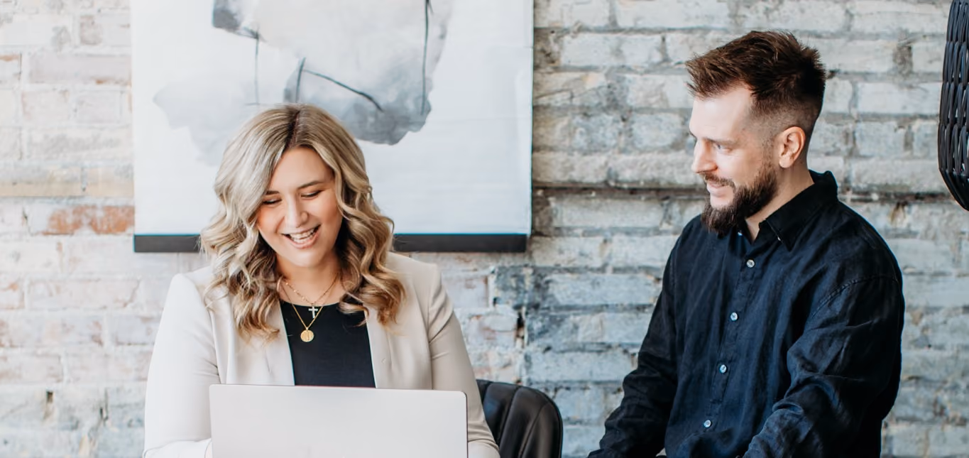 Smiling woman with blonde hair at laptop, man with beard watches, both seated against a brick wall.
