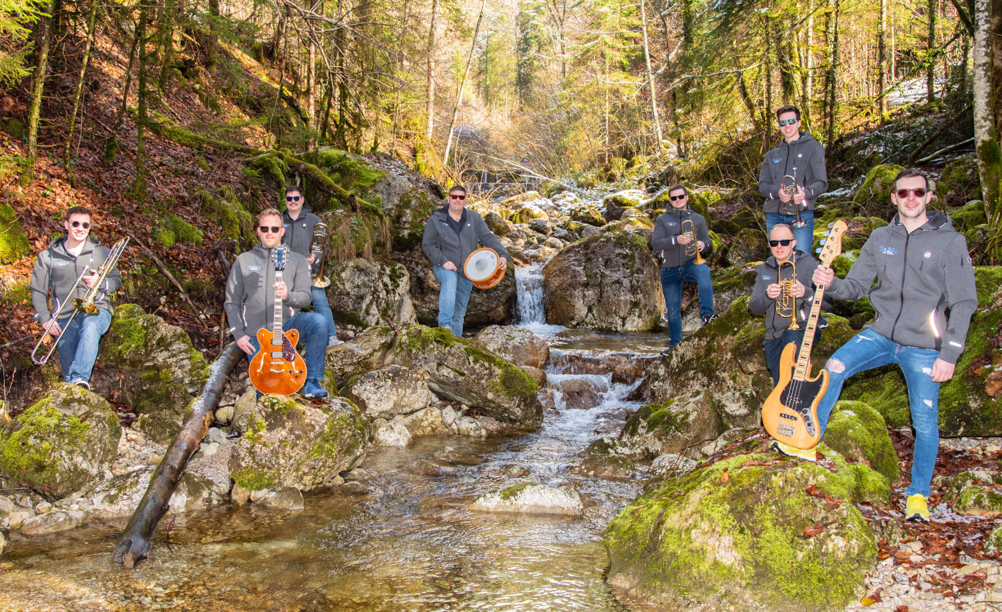 Eine Musikgruppe verteilt sich in einer idyllischen Waldszene auf mit Moos bewachsenen Felsen entlang eines klaren, sprudelnden Baches. Die Musiker tragen graue Kapuzenjacken, Sonnenbrillen und halten verschiedene Instrumente wie Trompeten, Posaunen, eine Gitarre, eine Bassgitarre und eine Trommel. Die Szene wirkt natürlich, mit sattem Grün und fließendem Wasser, das durch das Bild verläuft.