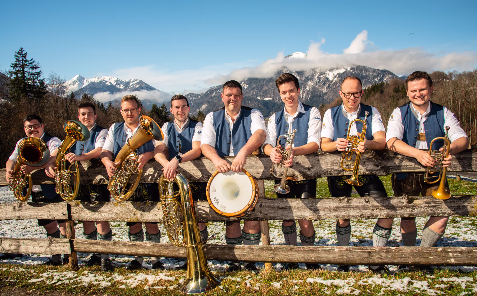 Eine Gruppe von acht Männern in traditioneller bayrischer Tracht steht hinter einem Holzzaun vor einer malerischen Alpenlandschaft. Sie halten verschiedene Blasinstrumente wie Trompeten, Tuben und ein Schlaginstrument. Im Hintergrund sind schneebedeckte Berge und ein blauer Himmel mit wenigen Wolken zu sehen. Der Boden ist teils mit Schnee bedeckt.