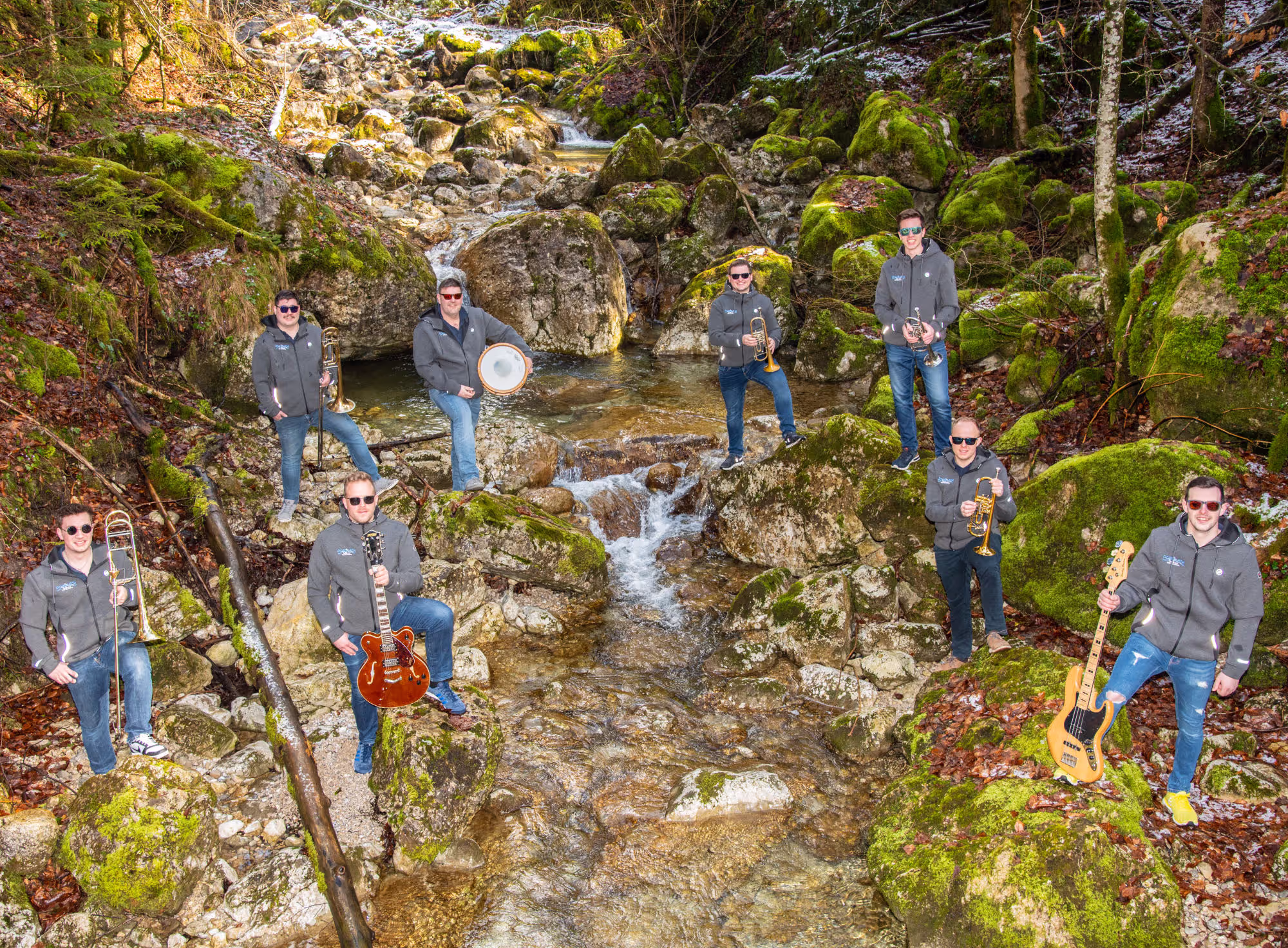 Eine Musikgruppe verteilt sich in einer idyllischen Waldszene auf mit Moos bewachsenen Felsen entlang eines klaren, sprudelnden Baches. Die Musiker tragen graue Kapuzenjacken, Sonnenbrillen und halten verschiedene Instrumente wie Trompeten, Posaunen, eine Gitarre, eine Bassgitarre und eine Trommel. Die Szene wirkt natürlich, mit sattem Grün und fließendem Wasser, das durch das Bild verläuft.