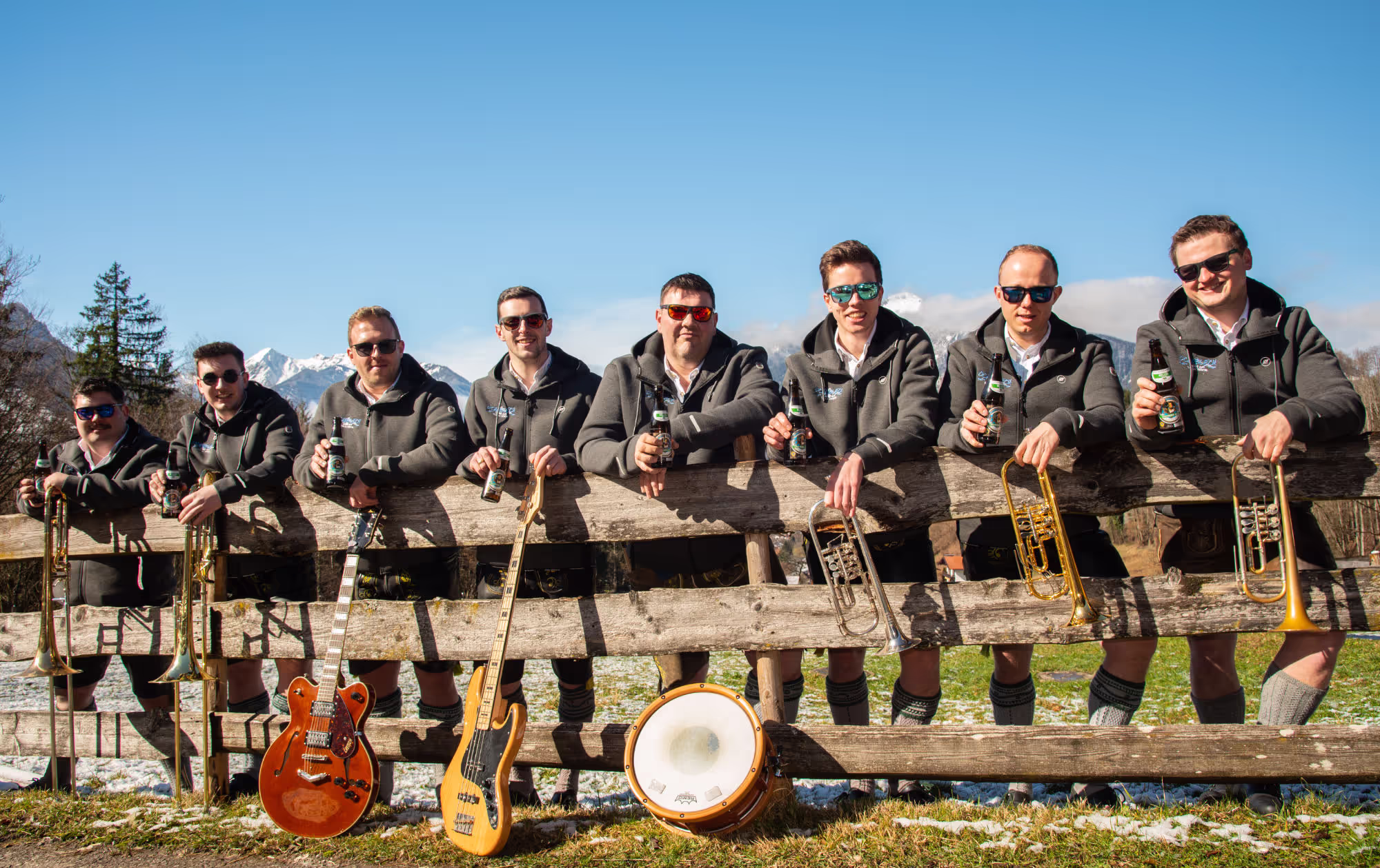 Eine Gruppe von neun Männern steht hinter einem Holzzaun in einer ländlichen, alpinen Umgebung. Sie tragen traditionelle Trachten (Lederhosen) und einheitliche graue Jacken. Jeder von ihnen hält ein Bier in der Hand. Vor ihnen lehnen Musikinstrumente, darunter Trompeten, eine Gitarre, ein Bass und eine Trommel. Im Hintergrund sind schneebedeckte Berge und ein blauer Himmel zu sehen.