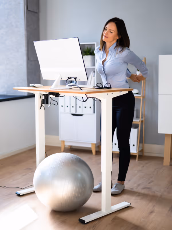 Woman in office stretching her back while standing at a height-adjustable desk with a silver exercise ball nearby.