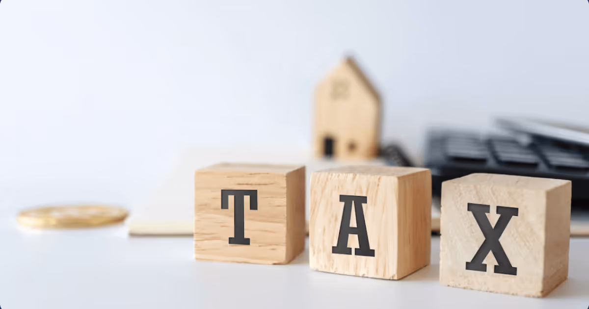 Three wooden blocks spelling 'TAX' with a blurred house model, calculator, documents, and coin in the background.