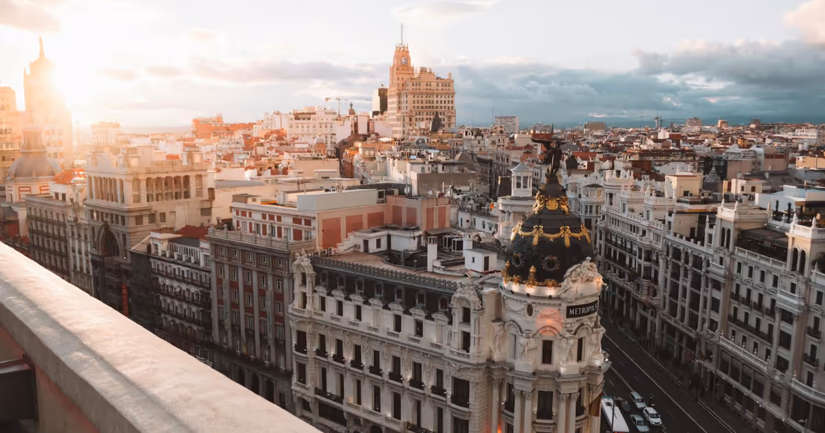 Cityscape of Madrid, Spain, featuring the iconic Metropolis Building with its ornate dome and golden details, surrounded by a mix of historic and modern architecture under a warm sunset glow.