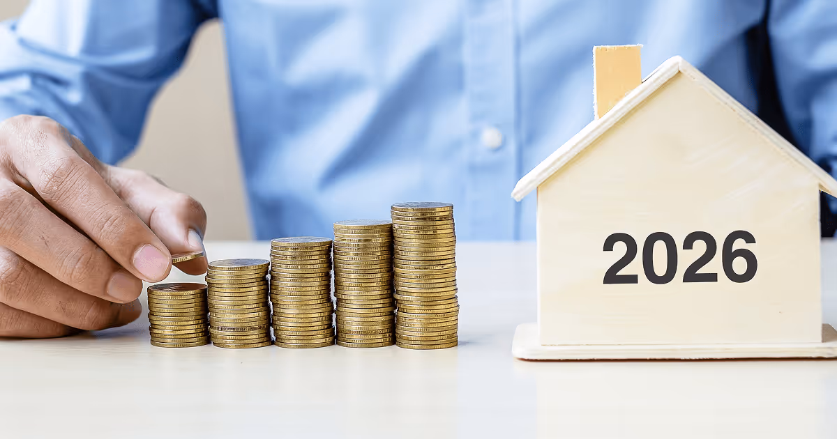 Hand placing coins on ascending stacks beside a wooden house model labeled 2026