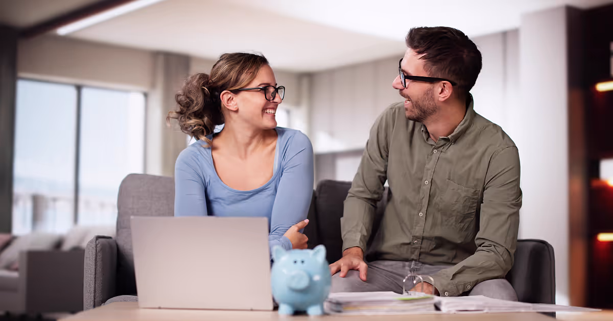 Two people smiling on a couch with a piggy bank, laptop, and papers on the coffee table.