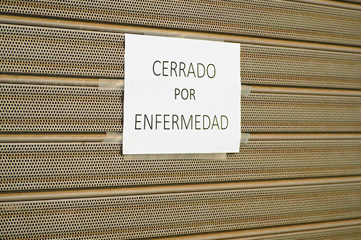 Metal shutter with paper sign reading “CERRADO POR ENFERMEDAD” indicating temporary closure due to illness