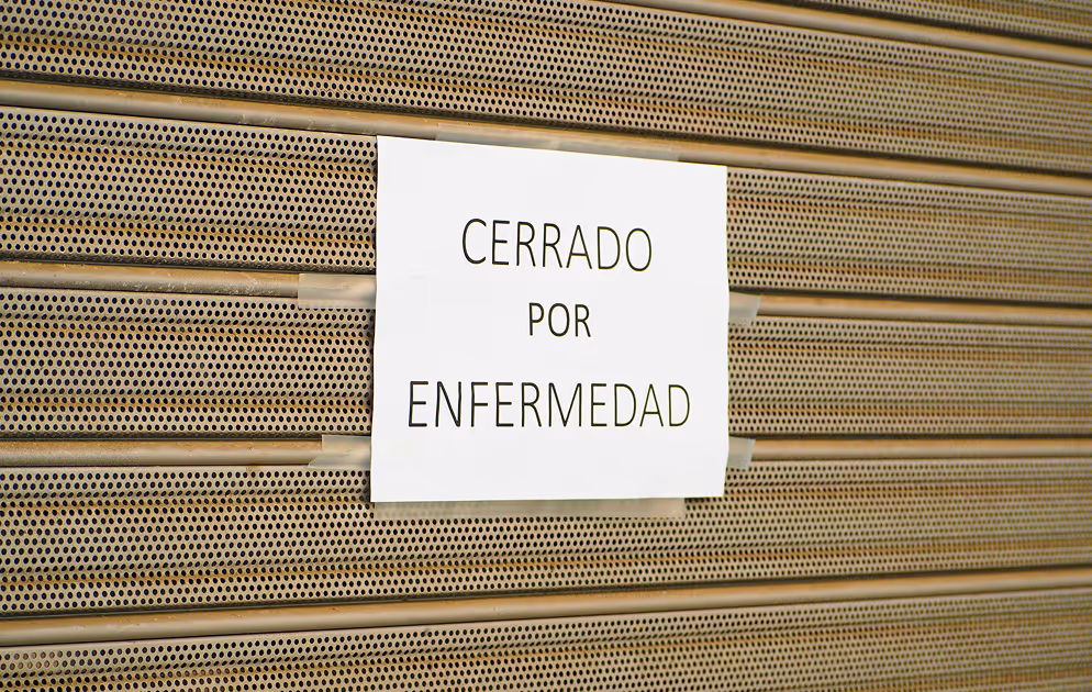 Metal shutter with paper sign reading “CERRADO POR ENFERMEDAD” indicating temporary closure due to illness