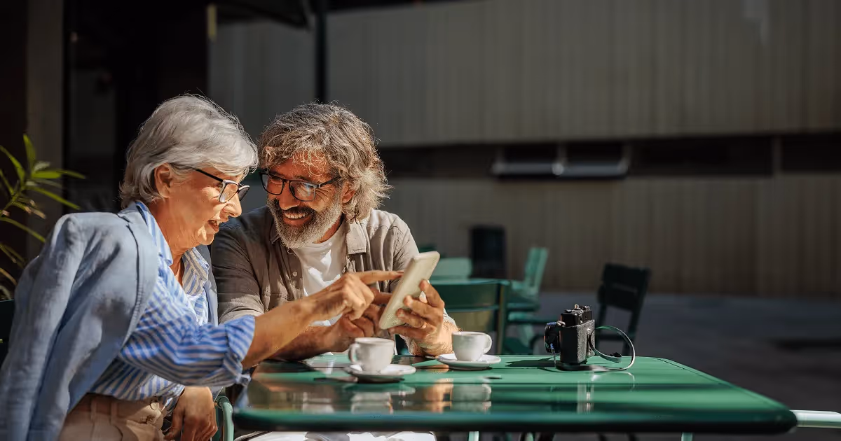 Smiling retired couple sitting at an outdoor café, enjoying coffee while looking at a tablet together.