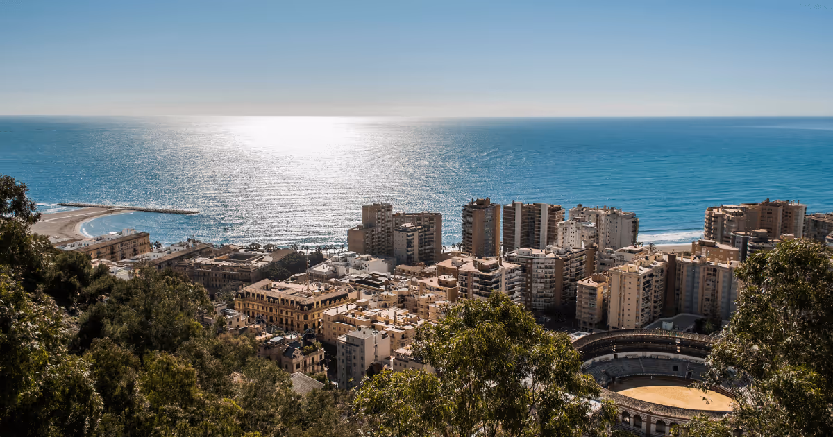 Elevated view of Málaga, Spain, showing coastal city buildings, the Mediterranean Sea, beachfront, and a historic bullring in the foreground under clear skies.