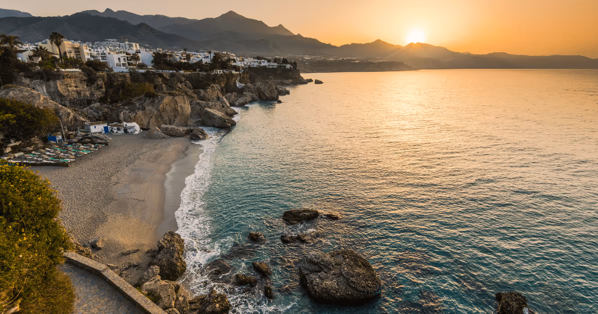 Golden sunrise over the Mediterranean Sea in Nerja, Andalusia, with rocky cliffs, a quiet beach, white coastal buildings, and mountains silhouetted in the background.