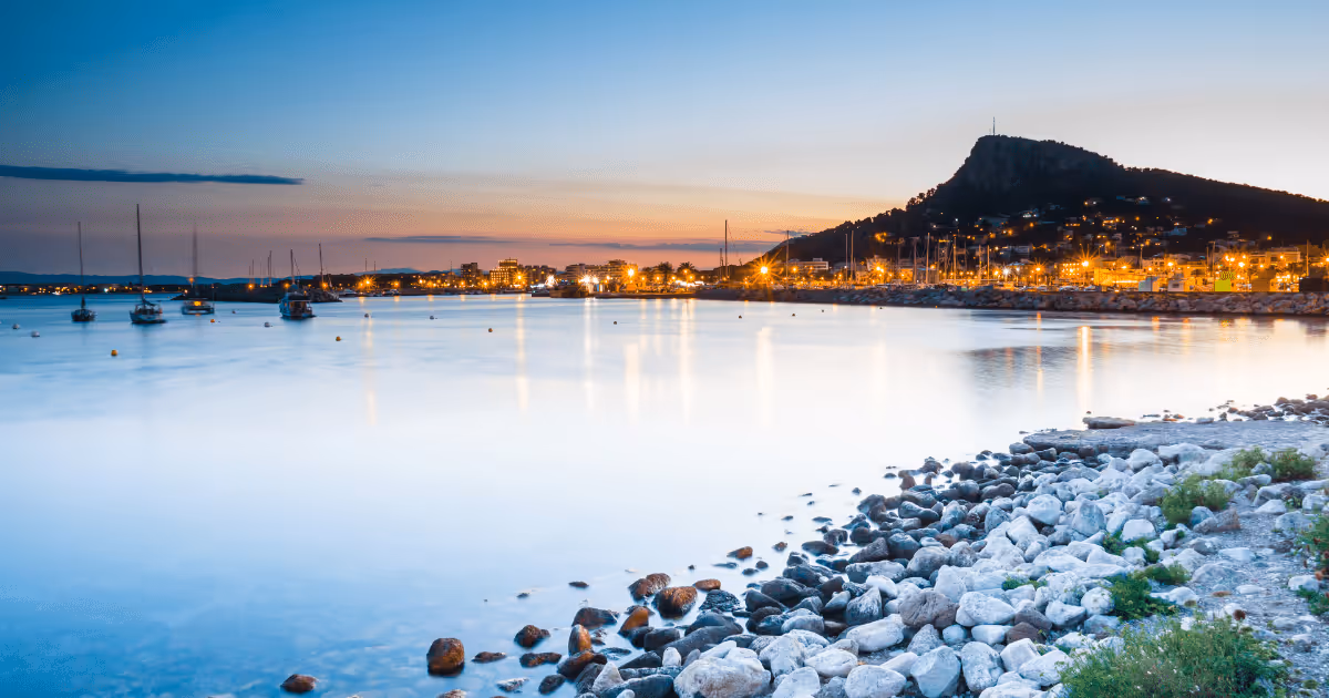 Sunset view of the harbor in L’Escala, Spain, with calm sea water, anchored sailboats, rocky shoreline in the foreground, and town lights reflecting on the sea along the Costa Brava