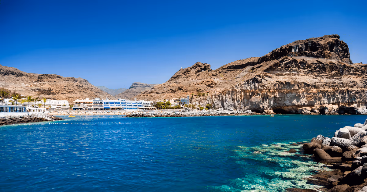 Beach on Gran Canaria in the Canary Islands, Spain, with clear turquoise water, rocky cliffs, seaside buildings, and dry mountain landscape under a deep blue sky