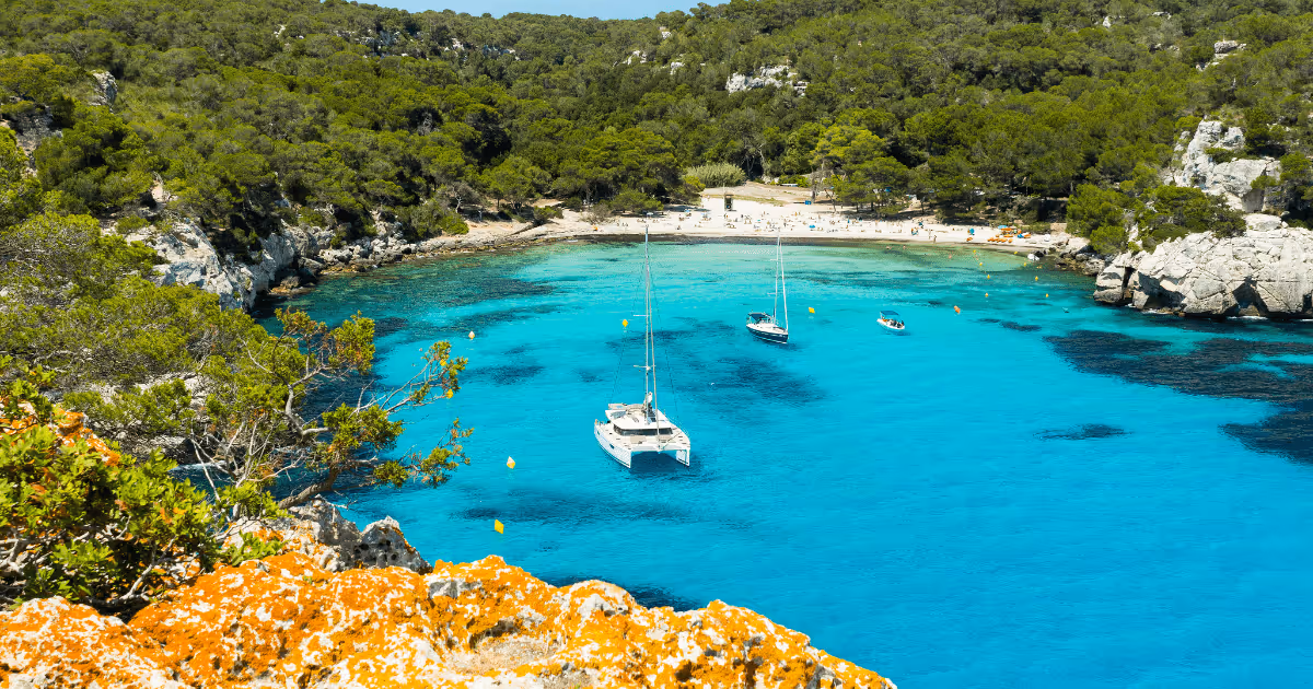 Cala Macarella in Menorca, Spain, with clear blue water, anchored sailboats, sandy beach, rocky cliffs, and pine forest surrounding the bay.