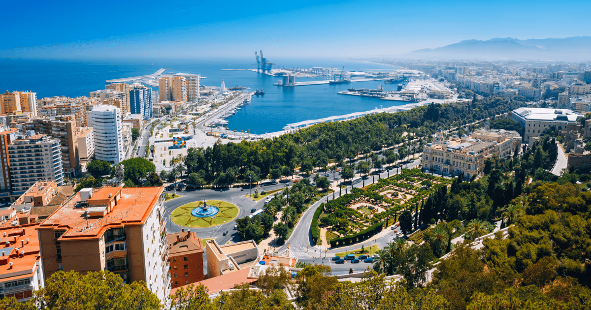 Aerial view of Málaga, Spain, showing the coastal cityscape, harbor and port area, green parks, roundabout, and the Mediterranean Sea under a clear blue sky.