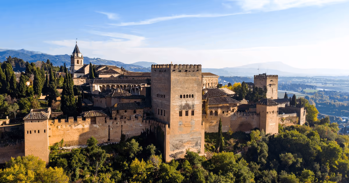 Panoramic view of the Alhambra palace complex in Granada, Spain, showing historic Moorish towers and walls surrounded by greenery with mountains in the background.