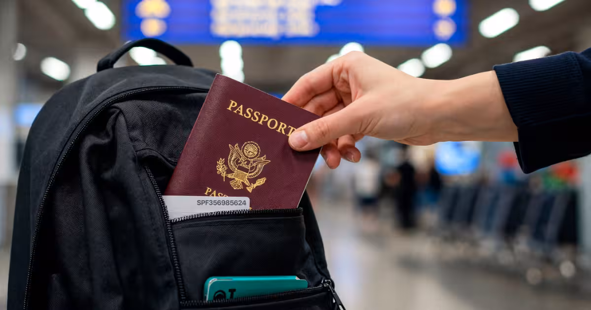Hand placing a passport into a backpack pocket inside an airport terminal, with departure boards blurred in the background, representing international travel and airport security.