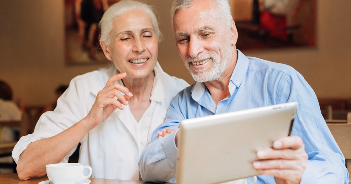 A smiling senior couple sitting at a table in a café, looking at a tablet together with cups of coffee in front of them.