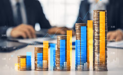 Stacks of coins arranged from low to high with an overlaid bar-and-line growth chart, while two business professionals sit blurred in the background at a desk.