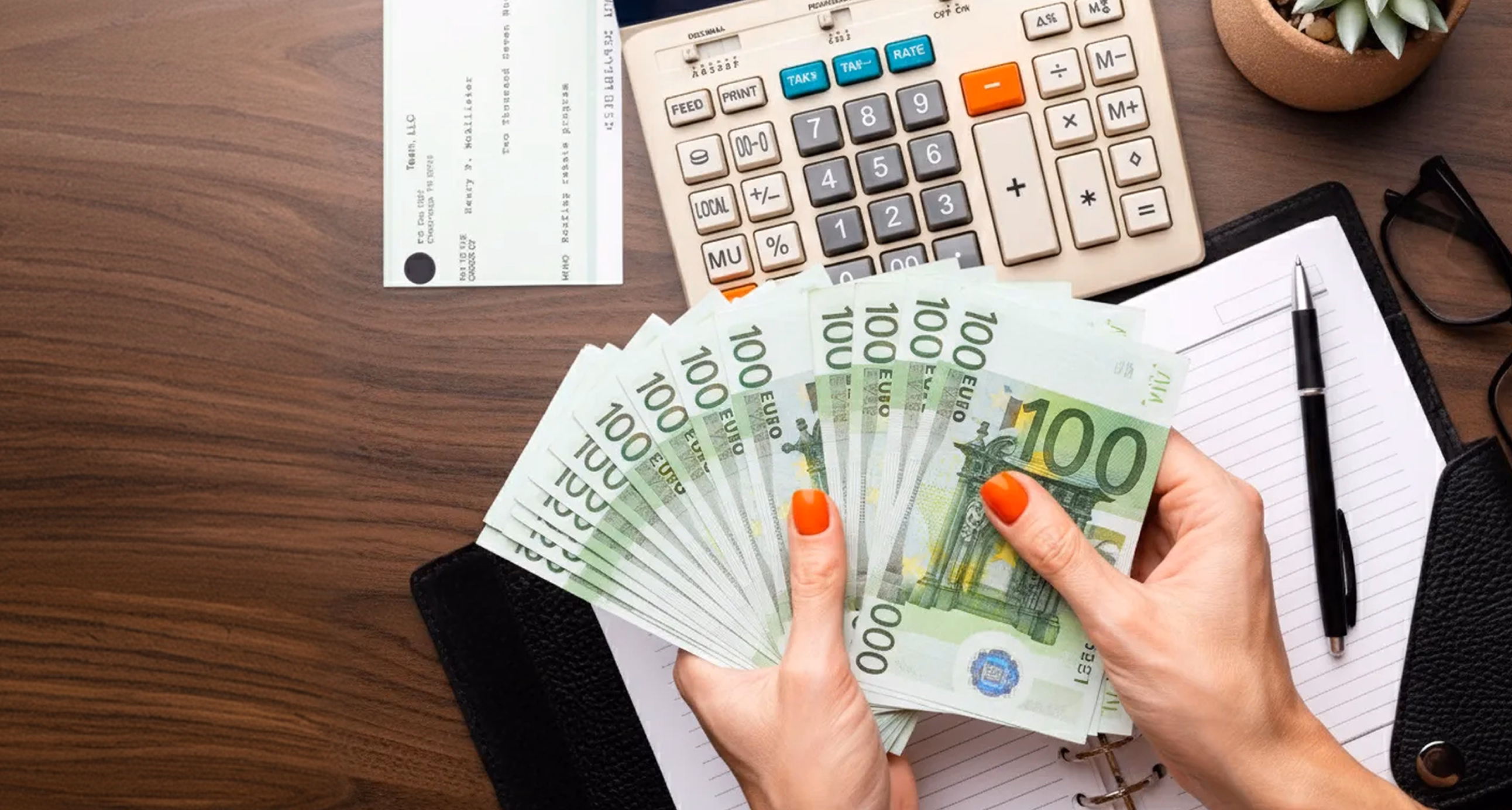 Hands holding €100 euro banknotes on a desk with a calculator, check, notebook, and pen.