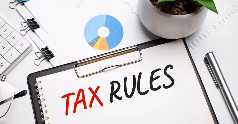 Notewith 'Tax Rules' written, placed on a clipboard surrounded by financial documents, a calculator, eyeglasses, pen, and a potted plant.