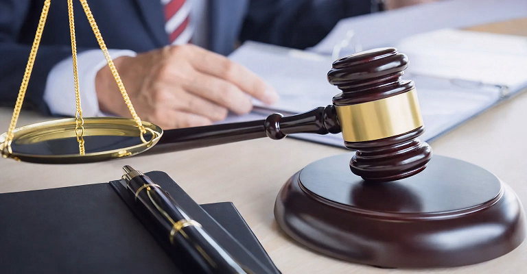 Close-up of a wooden judge’s gavel and gold balance scales on a desk with documents and a suited professional in the background, representing law and legal proceedings.