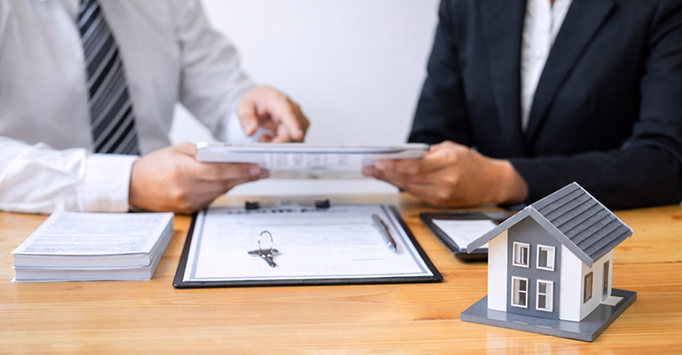 Two professionals reviewing property documents on a tablet at a wooden table with a clipboard and a small model house
