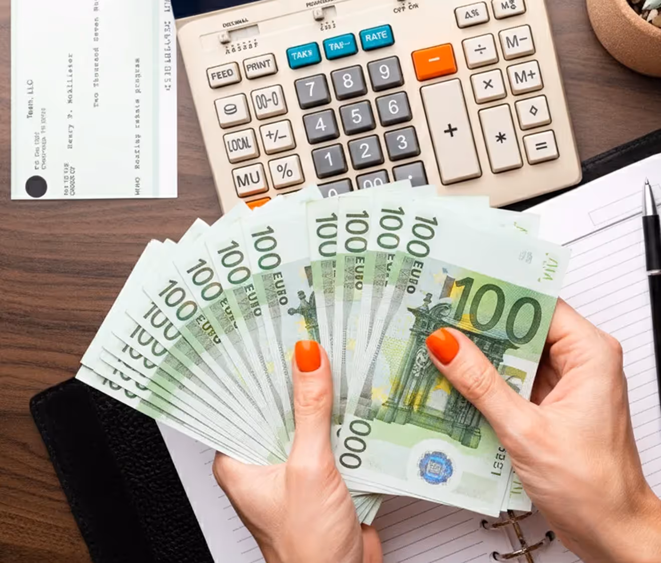 Hands holding €100 euro banknotes on a desk with a calculator, check, notebook, and pen.