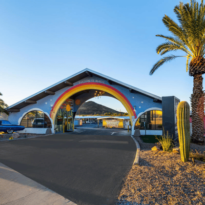 Entrance of the Hotel McCoy featuring a distinctive A-frame structure painted with a rainbow, flanked by a vintage blue car, palm trees, and tall cacti.